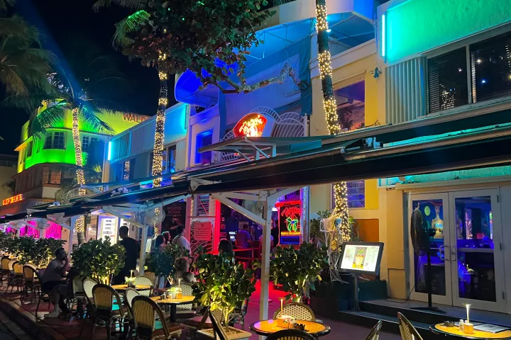 Mango's Tropical Cafe Miami Beach home of Salsa Mia Lessons, Classes, Nightlife Party, Live Music & Dancing. Nighttime street view with neon-lit outdoor seating and palm trees wrapped in lights in Miami, Florida