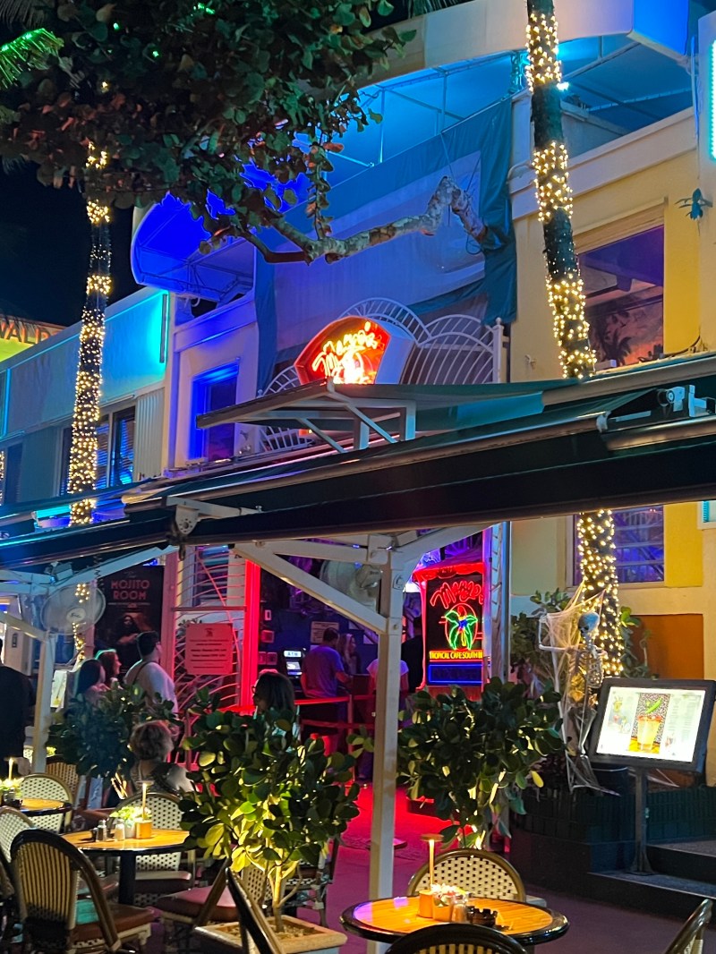 Mango's Tropical Cafe Miami Beach home of Salsa Mia Lessons, Classes, Nightlife Party, Live Music & Dancing. Nighttime street view with neon-lit outdoor seating and palm trees wrapped in lights in Miami, Florida