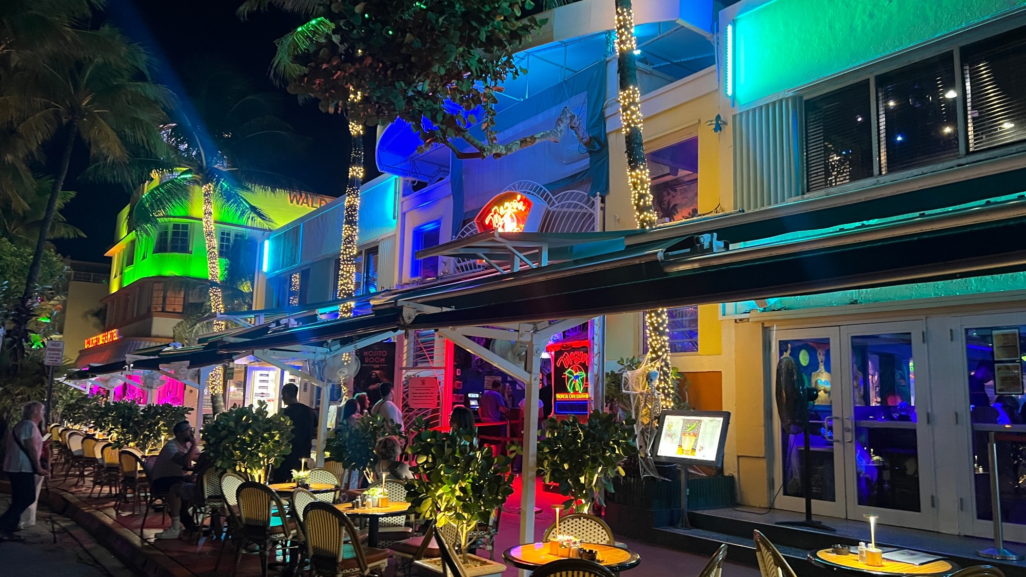 Mango's Tropical Cafe Miami Beach home of Salsa Mia Lessons, Classes, Nightlife Party, Live Music & Dancing. Nighttime street view with neon-lit outdoor seating and palm trees wrapped in lights in Miami, Florida